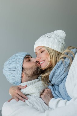 bearded man kissing cheek of happy blonde woman in knitted hat isolated on grey