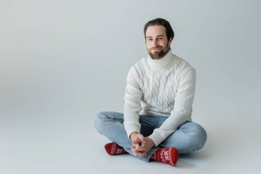 full length of bearded man in jeans and knitted sweater sitting in red socks with ornament on grey 