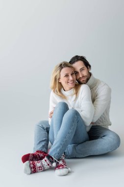 full length of happy couple in winter outfits and red socks with ornament sitting on grey