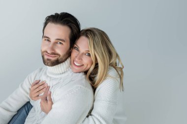 cheerful woman hugging bearded man in white sweater and looking at camera isolated on grey