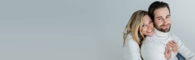cheerful woman hugging bearded man in white sweater and looking at camera isolated on grey, banner