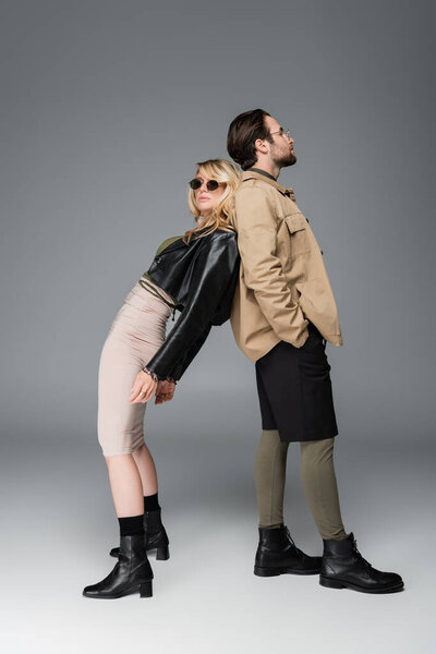 full length of stylish woman in trendy outfit and sunglasses leaning on man while posing on grey