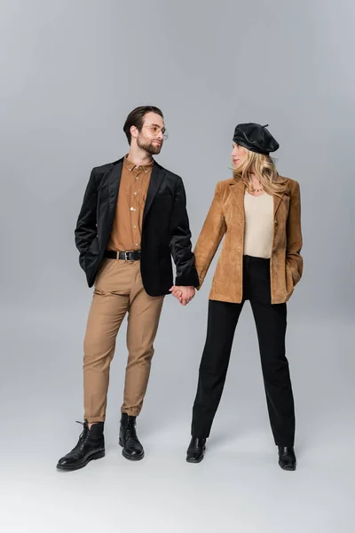 full length of blonde woman in stylish beret holding hands with bearded man in sunglasses on grey