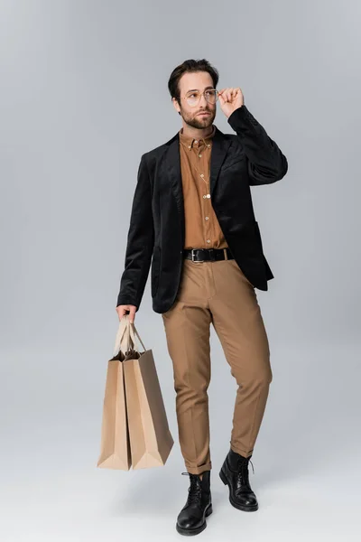 full length of bearded man in stylish autumnal outfit adjusting sunglasses and posing with shopping bags on grey