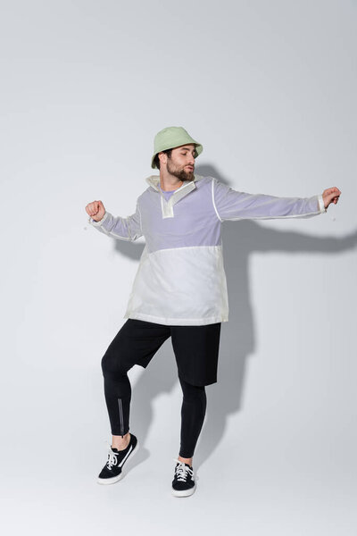 full length of bearded man in stylish sportive outfit and panama hat posing on grey