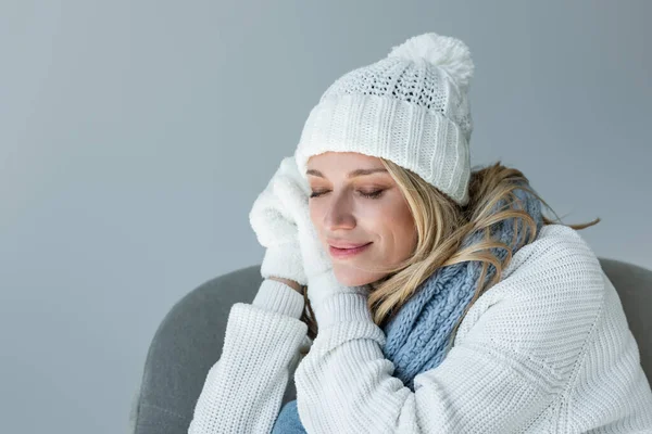 sleepy woman in winter outfit sitting in comfortable armchair and smiling isolated on grey