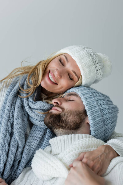 happy blonde woman in knitted scarf hugging with bearded boyfriend in winter hat isolated on grey