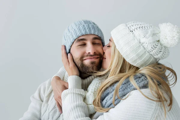 happy blonde woman in knitted hat kissing cheek of smiling boyfriend isolated on grey