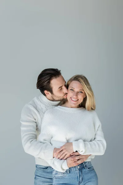 bearded man kissing cheek of blonde and smiling girlfriend isolated on grey