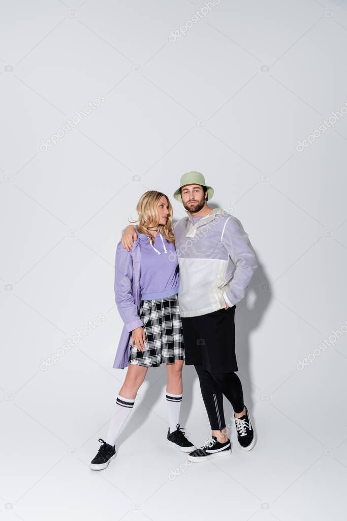 full length of man in panama hat hugging stylish woman in tartan skirt and longs socks with sneakers on grey