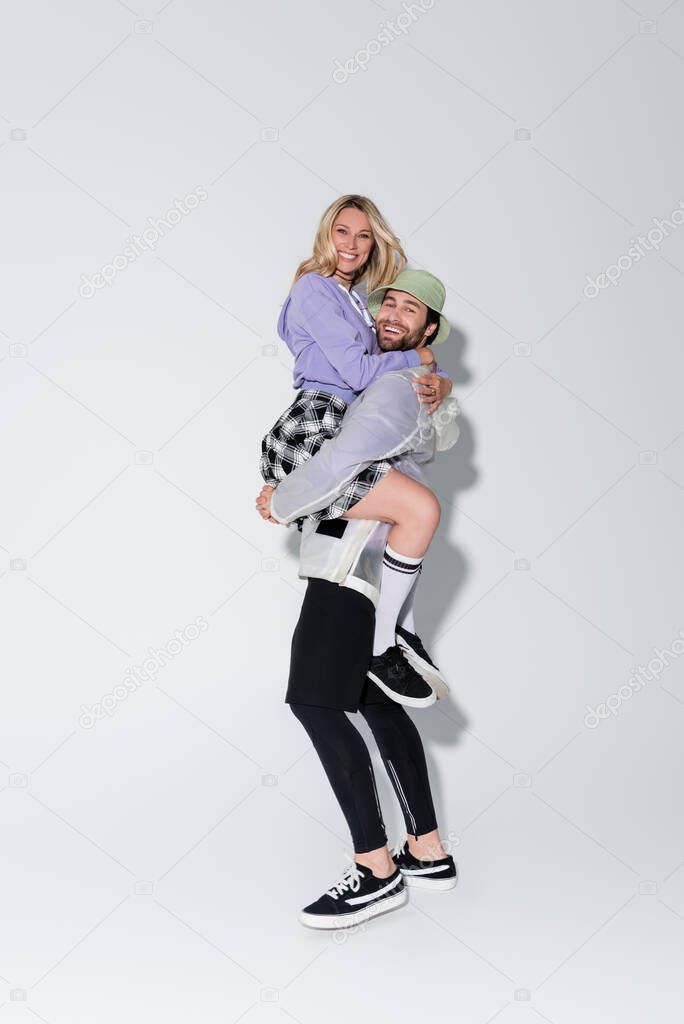 full length of happy man in panama hat holding in arms cheerful woman in tartan skirt and longs socks on grey