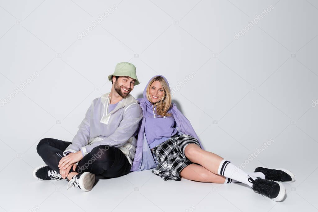full length of happy woman in tartan skirt and longs socks sitting with man in panama hat on grey