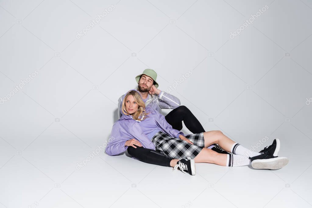 full length of man in stylish panama hat hugging blonde woman in tartan skirt and longs socks while sitting on grey