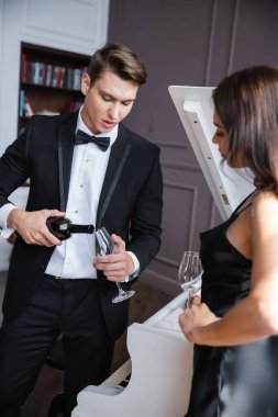 Elegant man pouring wine near blurred girlfriend and piano at home 