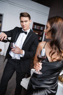 Elegant man pouring wine in glass near blurred girlfriend in dress at home 