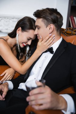 Cheerful woman with red lips touching boyfriend in suit holding blurred glass at home 