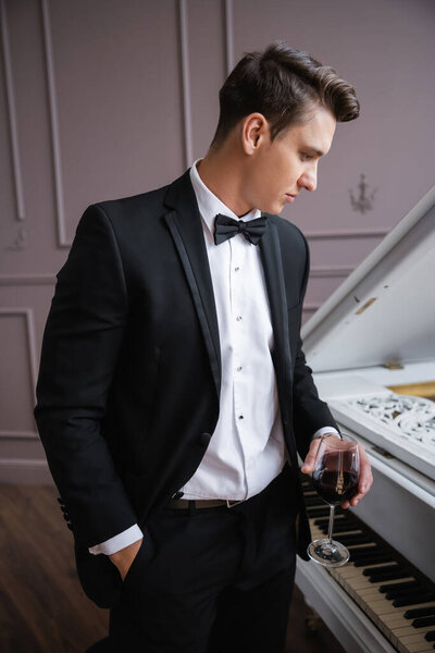 Side view of elegant man in suit holding glass of wine near piano at home 