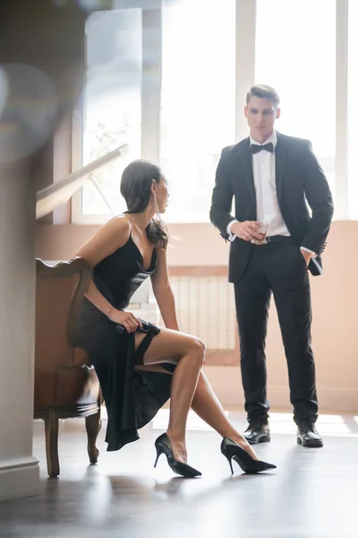 Sensual woman in heels and dress putting handgun in garter near boyfriend with whiskey at home