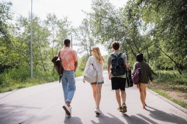 back view of multiethnic tourists with backpacks walking on road on summer day