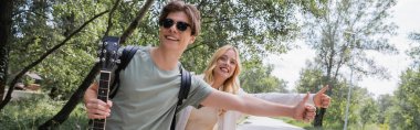 young and cheerful hitchhikers with thumbs up stopping car in countryside, banner