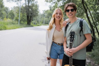 blonde woman leaning on man in sunglasses while looking at camera on road