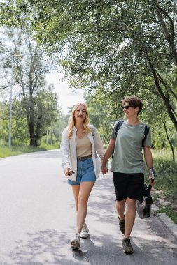 full length of young travelers holding hands and walking on road along forest