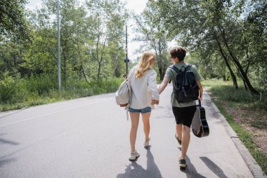 back view of young tourists with backpacks and guitar holding hands and walking on road