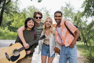 happy african american man standing with hand in pocket near multicultural friends with guitar and backpacks