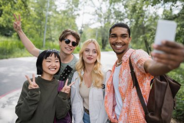 happy african american man taking selfie with interracial friends showing victory signs