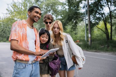 smiling african american man searching direction on digital tablet near multiethnic friends