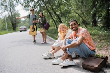 blurred hitchhikers stopping car near smiling interracial friends sitting on road