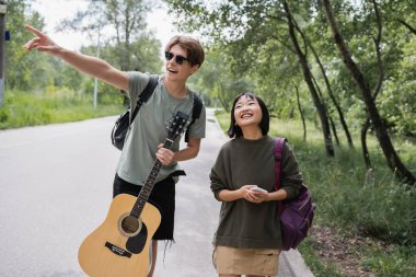 young man with guitar pointing with finger near cheerful asian girlfriend