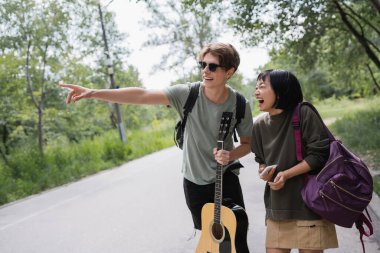 smiling man in sunglasses pointing with finger near laughing asian girlfriend