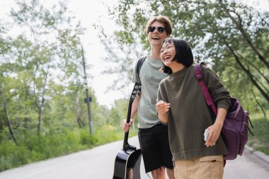 excited asian woman laughing near boyfriend in sunglasses while walking in countryside