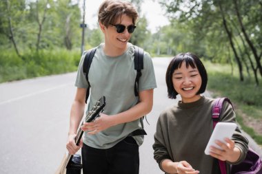 smiling asian woman searching direction on smartphone near young man in sunglasses