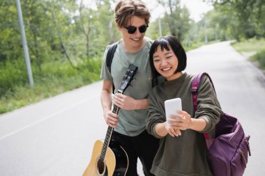 asian woman searching route on smartphone near boyfriend with acoustic guitar