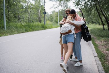 happy multiethnic travelers embracing on countryside road