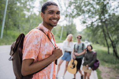 young african american man with backpack smiling at camera near friends on blurred background