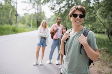 smiling man with sunglasses and backpack near multiethnic friends on blurred background