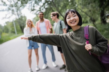 happy asian woman stopping car near young multiethnic friends on blurred background