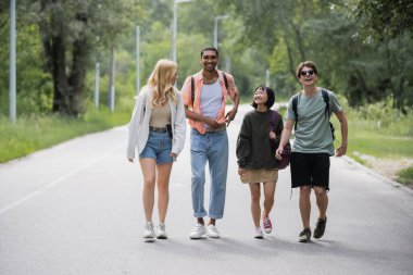 full length of young interracial travelers with backpacks walking along road near forest