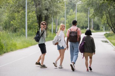 man in sunglasses pointing with finger near interracial friends walking on road along forest