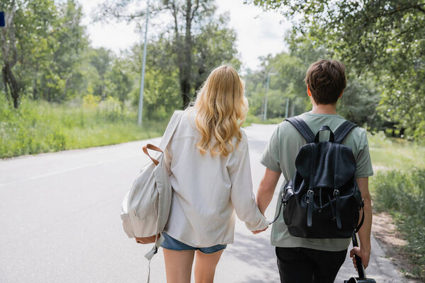 back view of young travelers holding hands and walking on road along forest