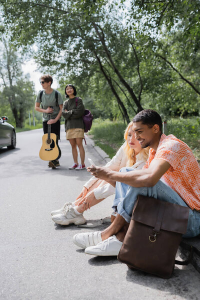 Африканский американец ищет маршрут на смартфоне, сидя на дороге рядом с межрасовыми друзьями