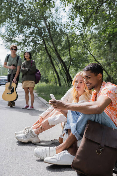 веселый африканский американец ищет направление на смартфоне, сидя на дороге рядом с многонациональными друзьями