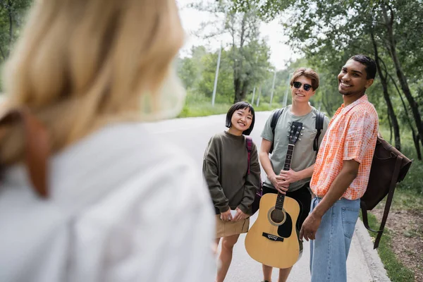 Happy Interracial Travelers Guitar Backpacks Looking Friend Blurred Foreground — Stockfoto