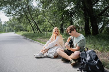 man playing guitar while sitting on road near blonde girlfriend