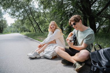 man sitting on road with crossed legs and playing guitar to blonde girlfriend