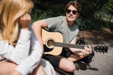 man in sunglasses sitting with crossed legs and playing guitar to blurred girlfriend