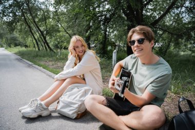 man playing guitar on road near happy blonde girlfriend